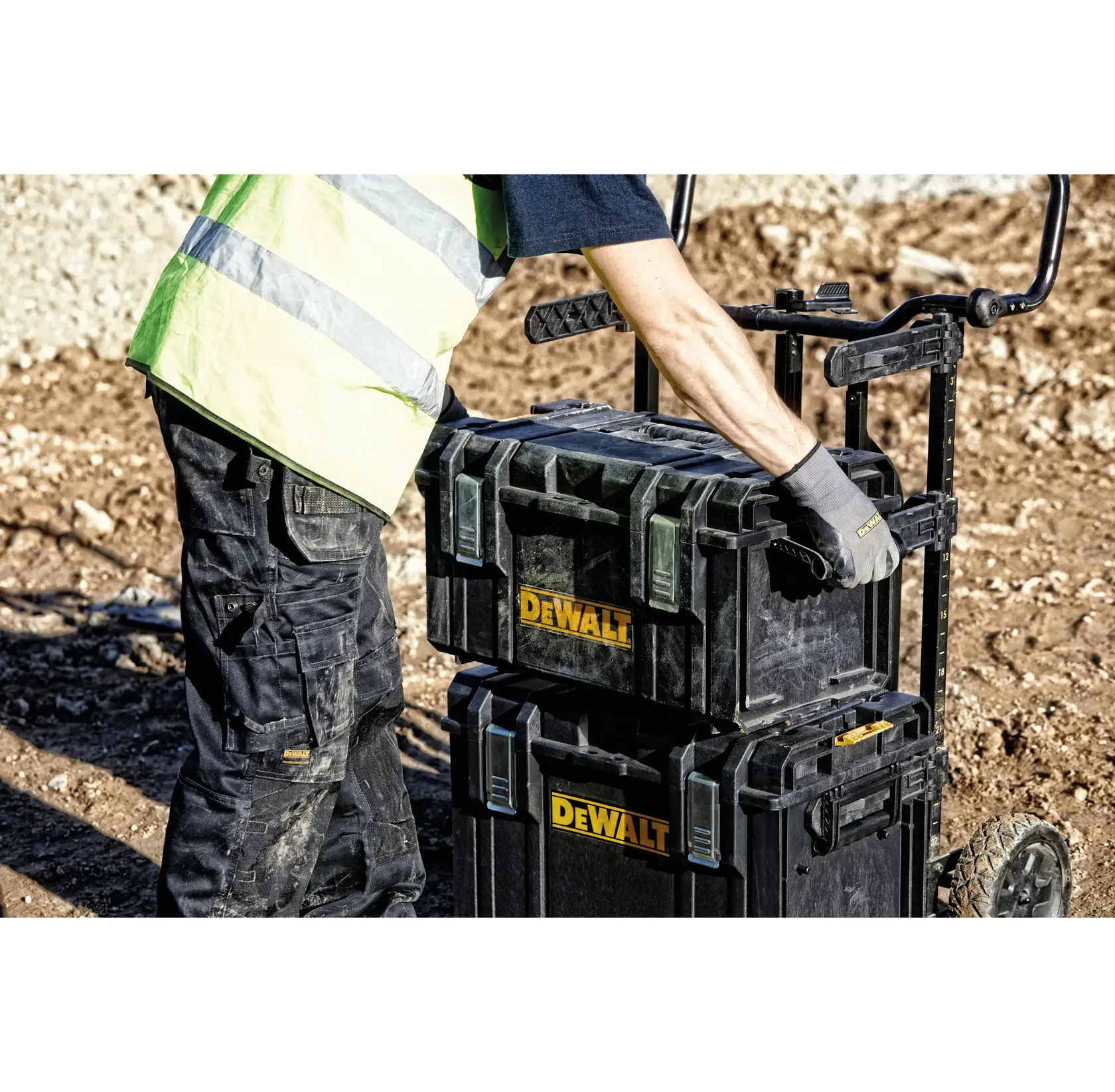 A person wearing a high-visibility vest and work gloves moves DEWALT toolboxes stacked on a trolley cart at a construction site.