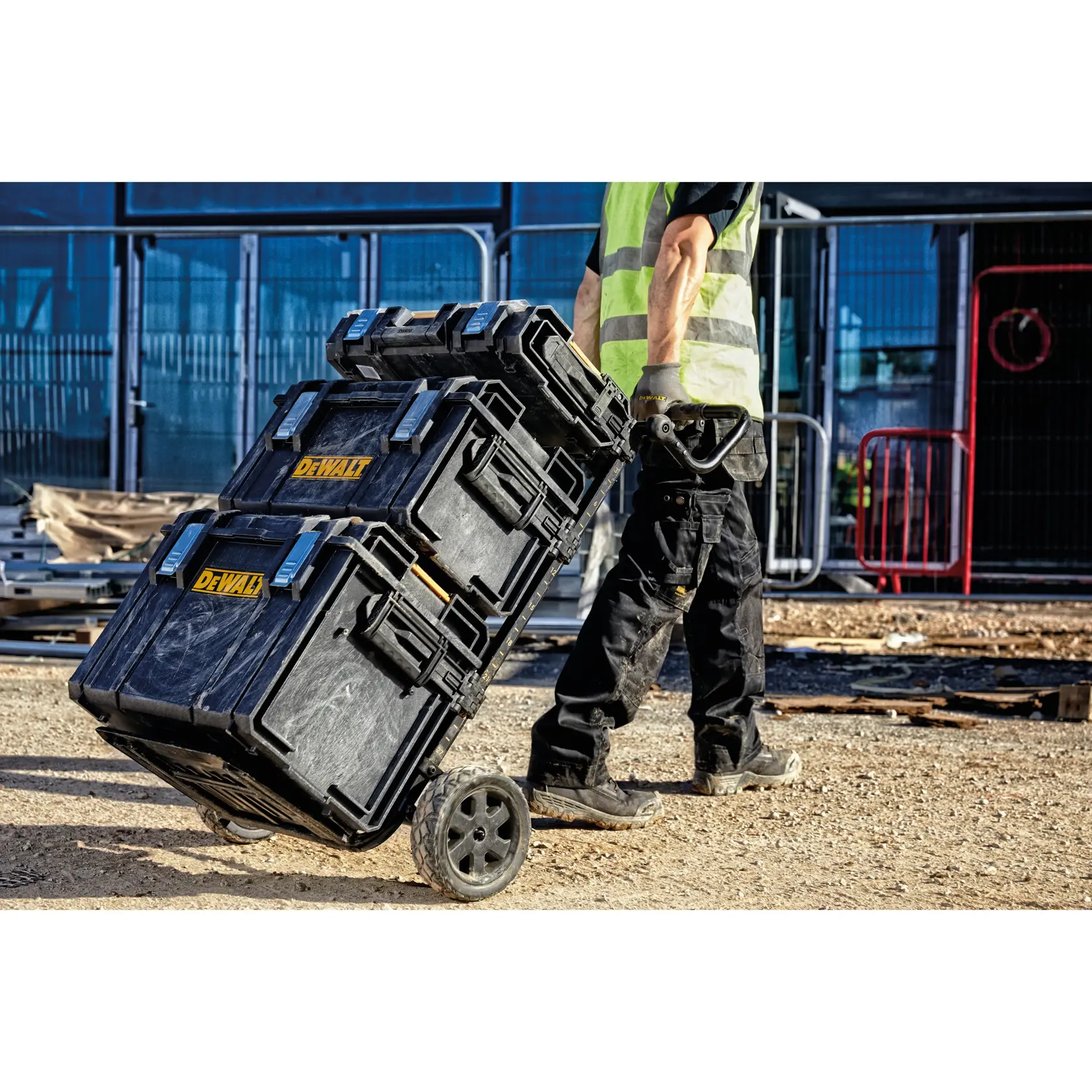 A person wearing work clothes and a high-visibility vest is pulling a stack of three DEWALT black toolboxes on a wheeled trolley at a construction site.