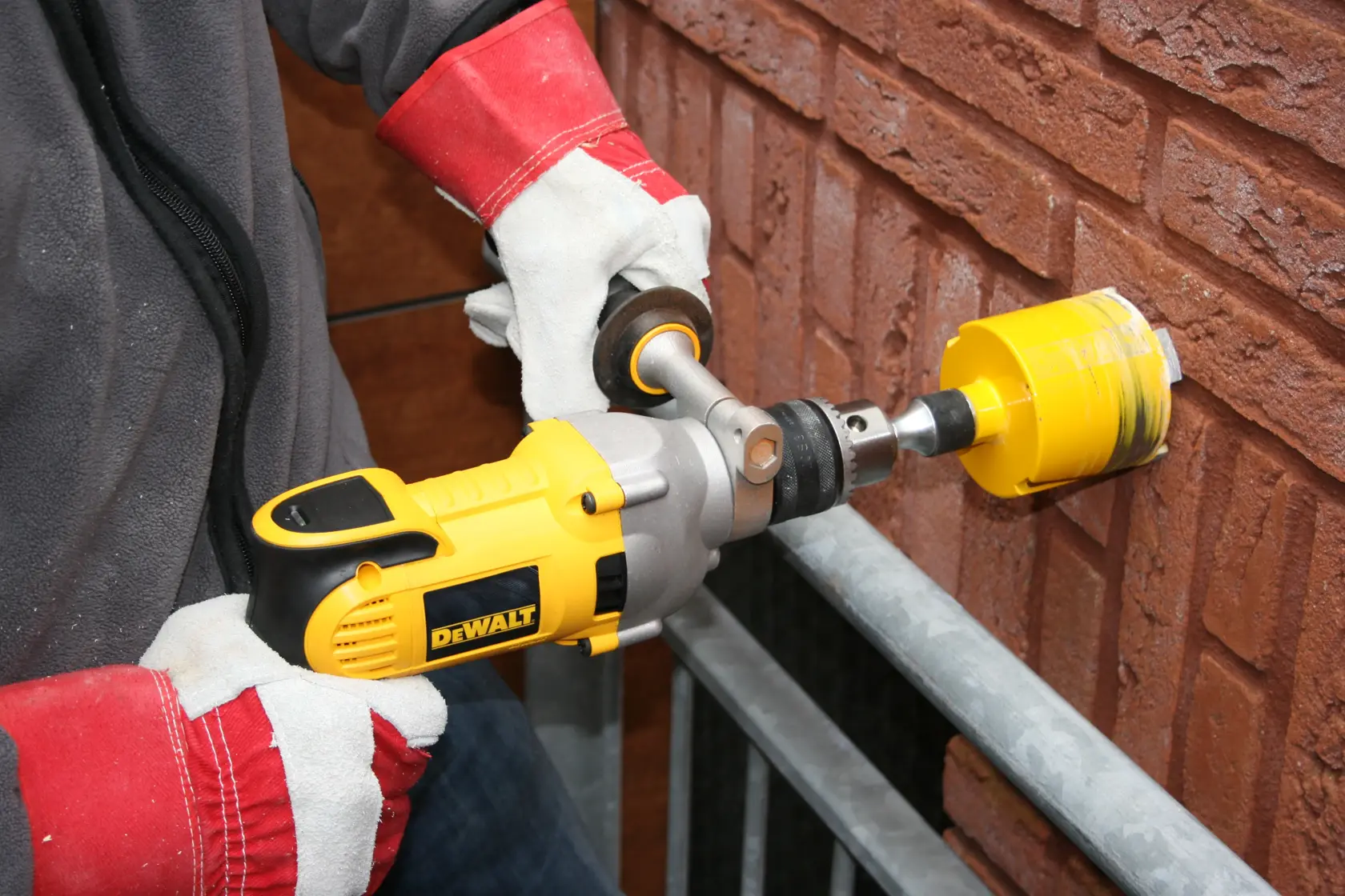 Una persona con guanti protettivi sta usando un trapano elettrico giallo DEWALT con una punta a fresa per forare un muro di mattoni.