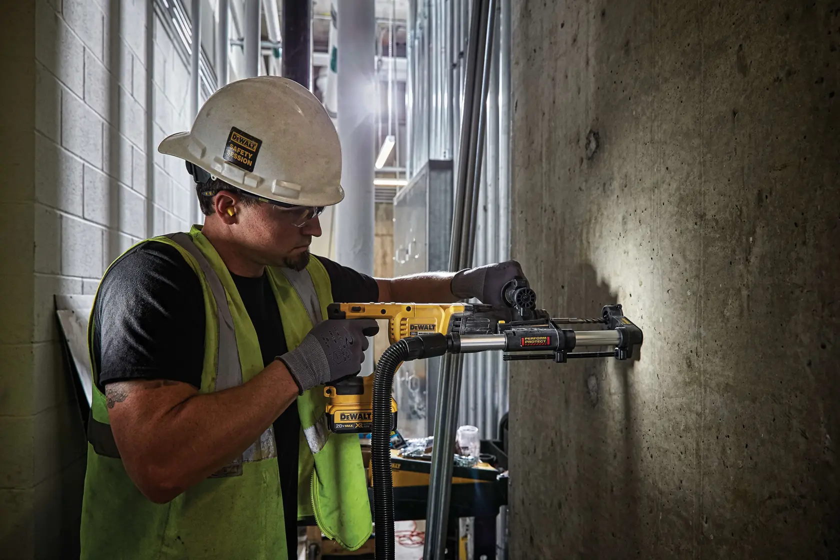 A construction worker wearing a hard hat and safety vest operates a yellow DEWALT power tool with a dust extraction attachment, drilling into a concrete wall in an industrial setting.