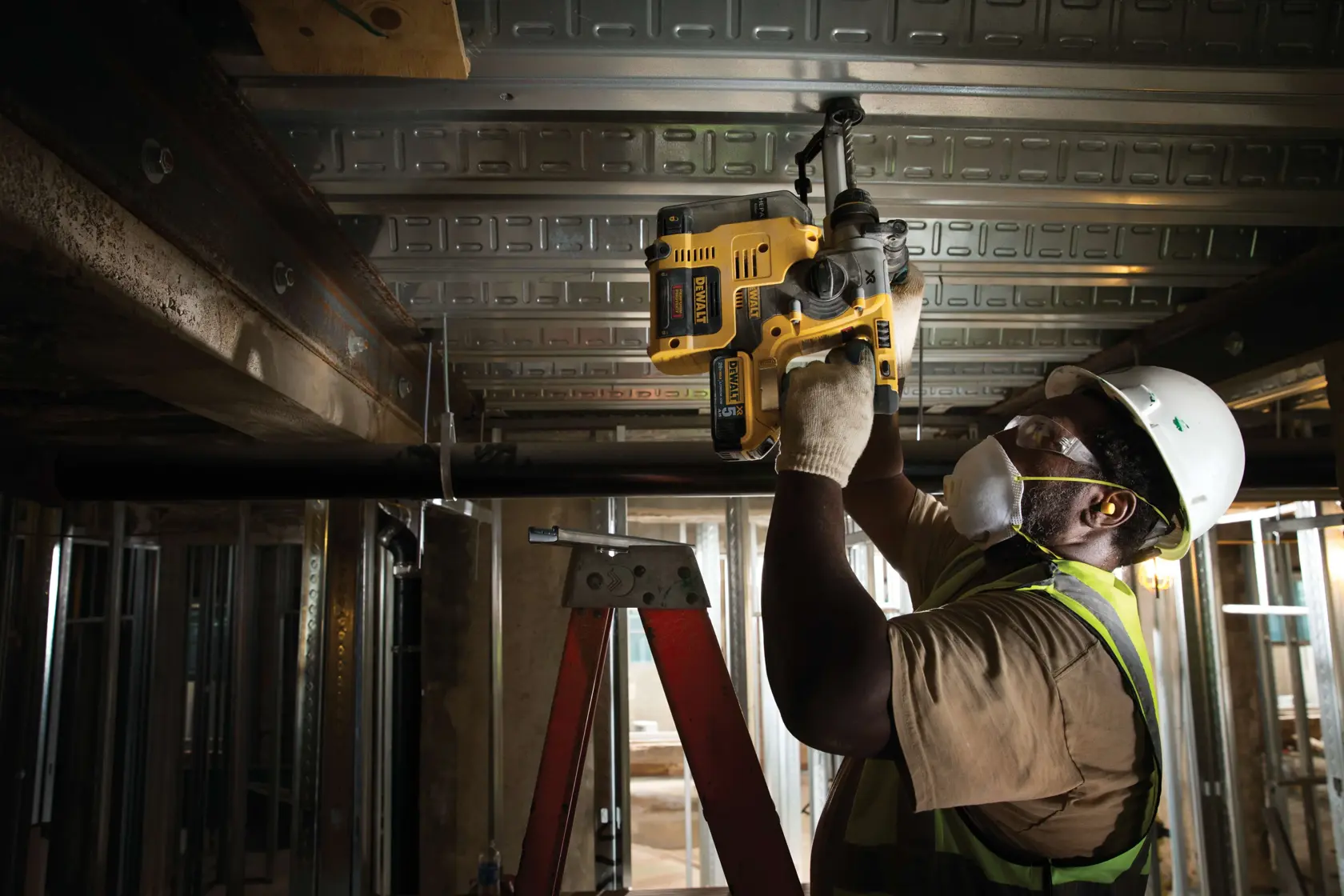 A construction worker wearing a hard hat, safety vest, gloves, and mask uses a DEWALT D25303DH tool to drill into a metal ceiling beam while standing on a ladder at a building site.
