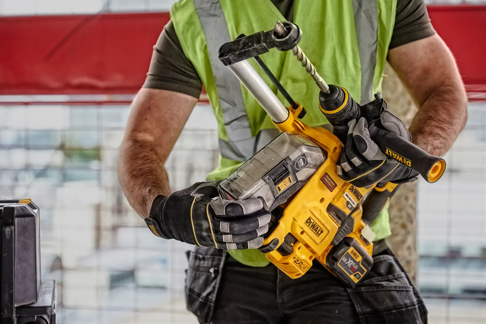 A person wearing a green safety vest and gloves is holding a DEWALT D25304DH cordless hammer drill with an attached dust extraction system on a construction site.