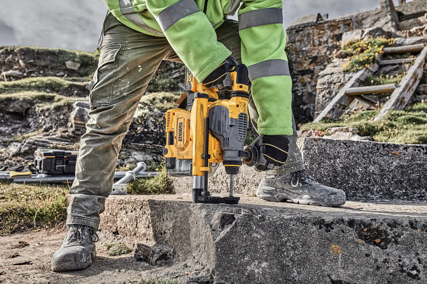 Person using DEWALT drill on concrete outdoors, wearing gloves, boots, and safety gear.