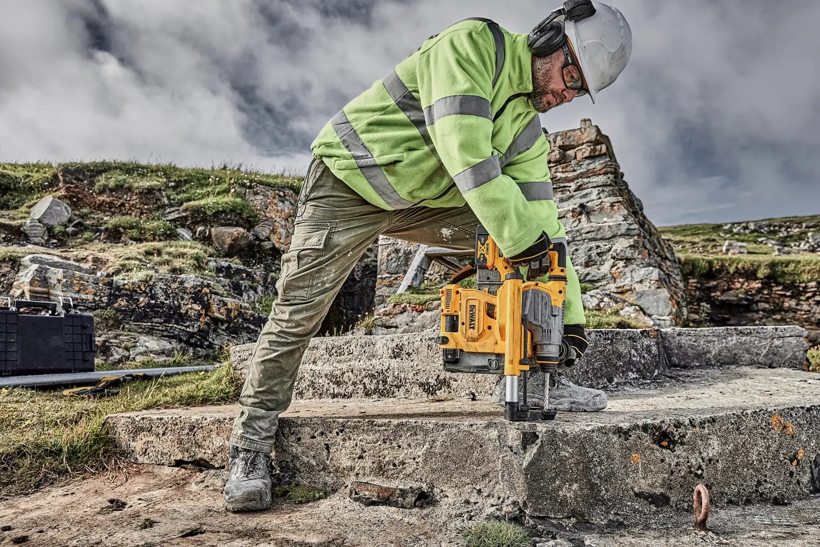 Worker drills into concrete outdoors using a yellow DEWALT tool, wearing safety gear and a high-visibility jacket.
