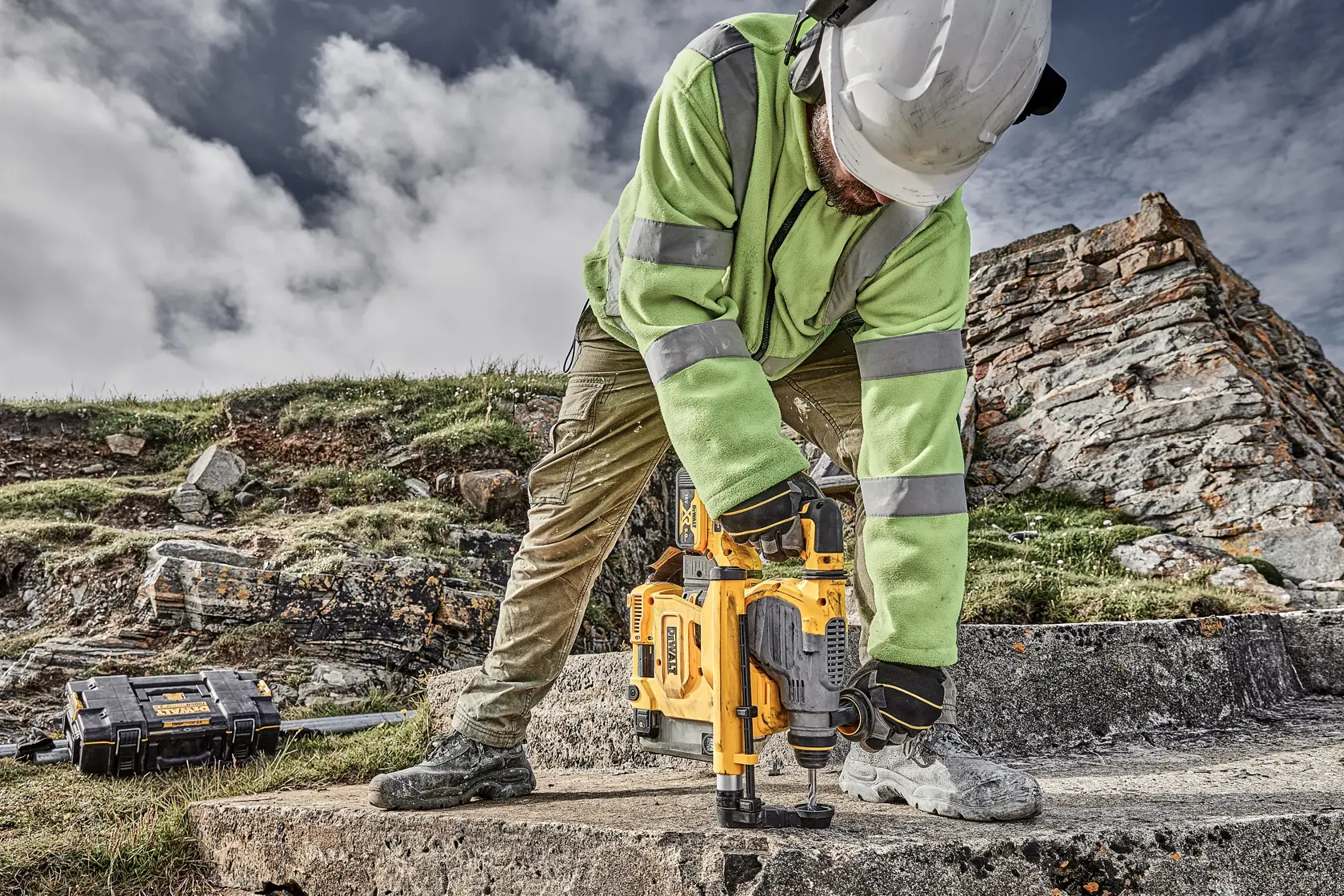 Worker using a DEWALT tool outdoors on concrete, wearing safety gear and a helmet, with a tool case nearby.