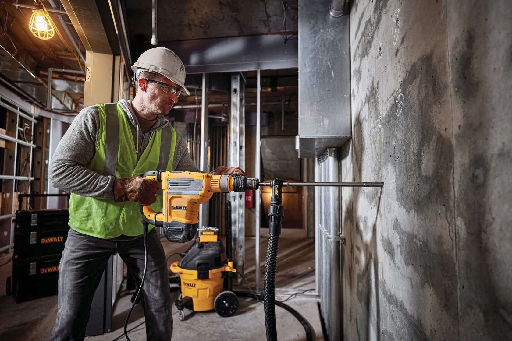 A person wearing safety gear and a helmet operates a DEWALT D25614K rotary hammer drill against a concrete wall in a construction site. DEWALT equipment and tools are visible in the background.