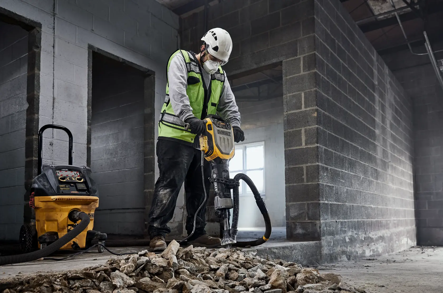 A construction worker wearing a safety vest and helmet is using a DEWALT jackhammer (model D25966) to break up concrete inside an unfinished building. There is a DEWALT dust extractor connected to the jackhammer, and broken concrete rubble is scattered on the floor.
