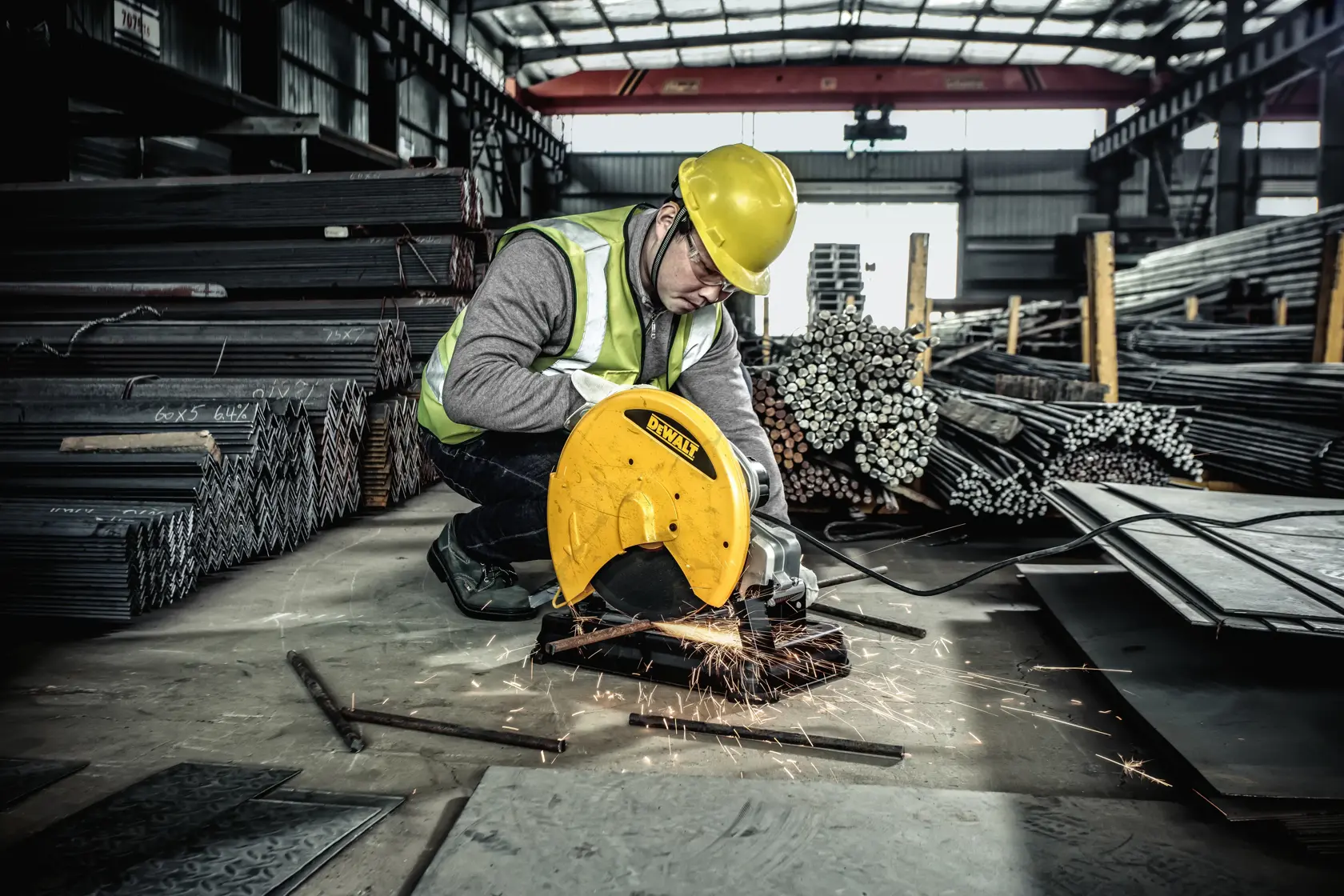A worker wearing a yellow safety helmet and reflective vest is cutting metal with a yellow DEWALT chop saw, producing sparks. The worker is in an industrial warehouse surrounded by stacks of metal bars and sheets.