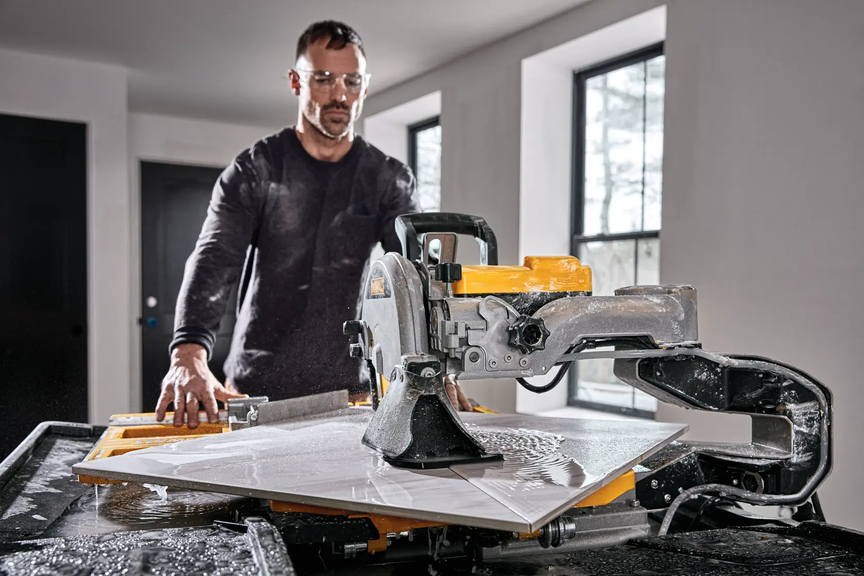 A person operating a DEWALT D36000-QS wet tile saw in a well-lit room, cutting a large tile with water splashing on the work surface.