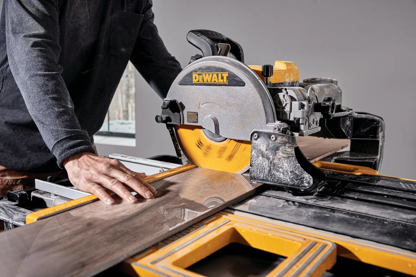 A person is cutting a large tile with a DEWALT tile saw. The saw blade is spinning and the person is guiding the tile carefully with their hand.