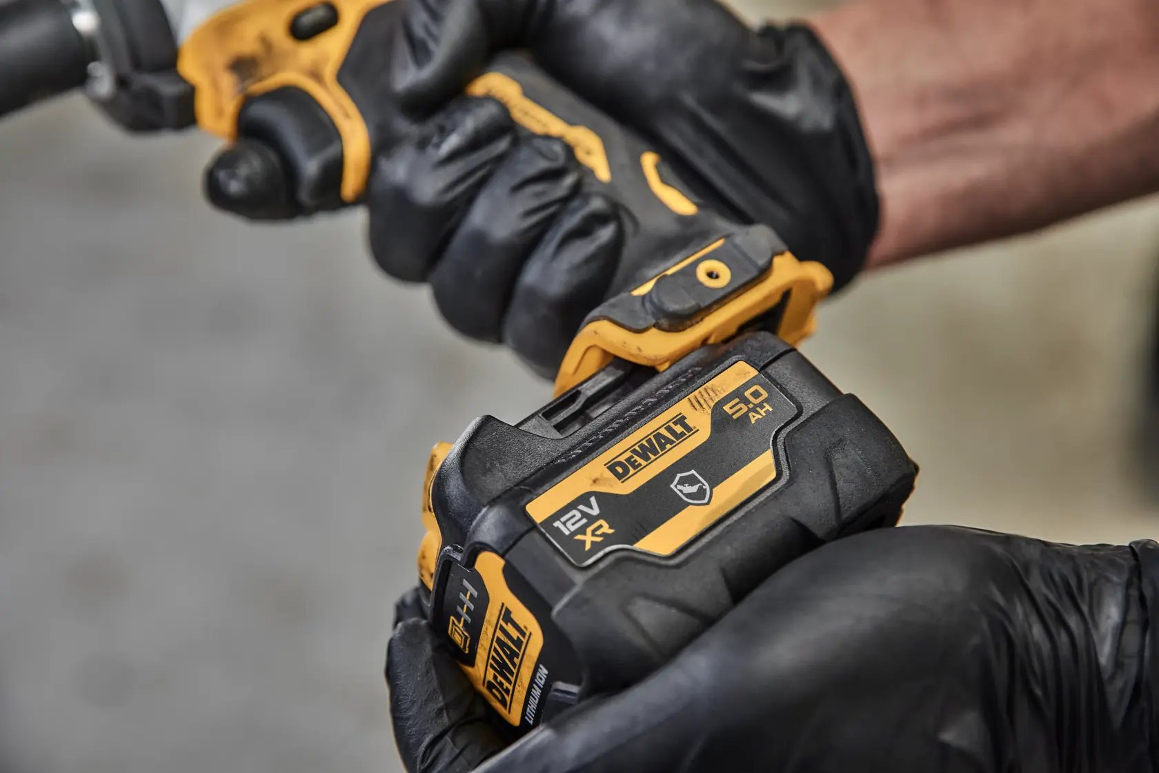Close-up image of a person wearing black gloves holding and installing a DEWALT 12V XR 5.0Ah battery, model DCB126G, onto a compatible tool.