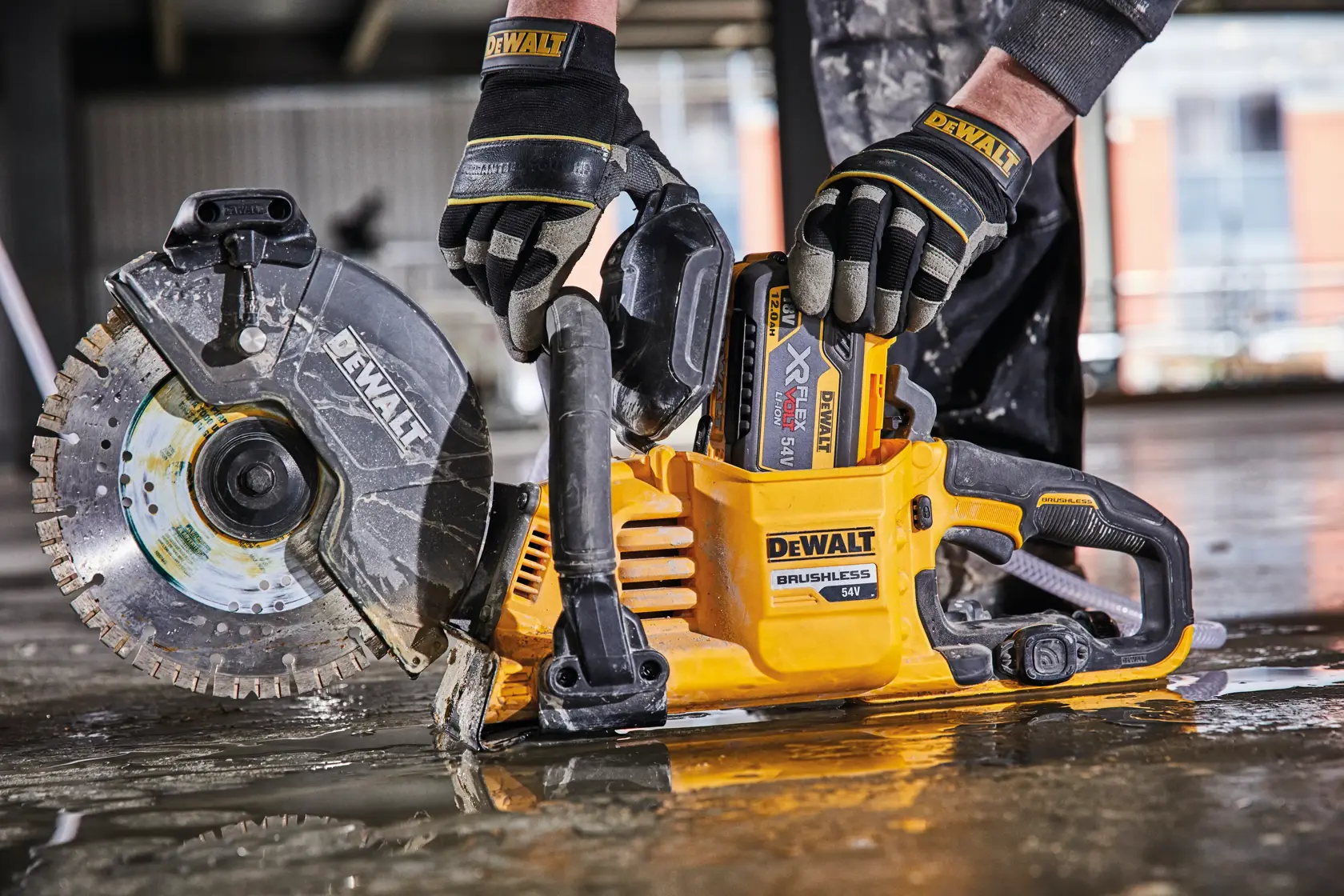 A person wearing protective gloves is handling a DEWALT 54V brushless cordless cut-off saw on a wet construction floor.