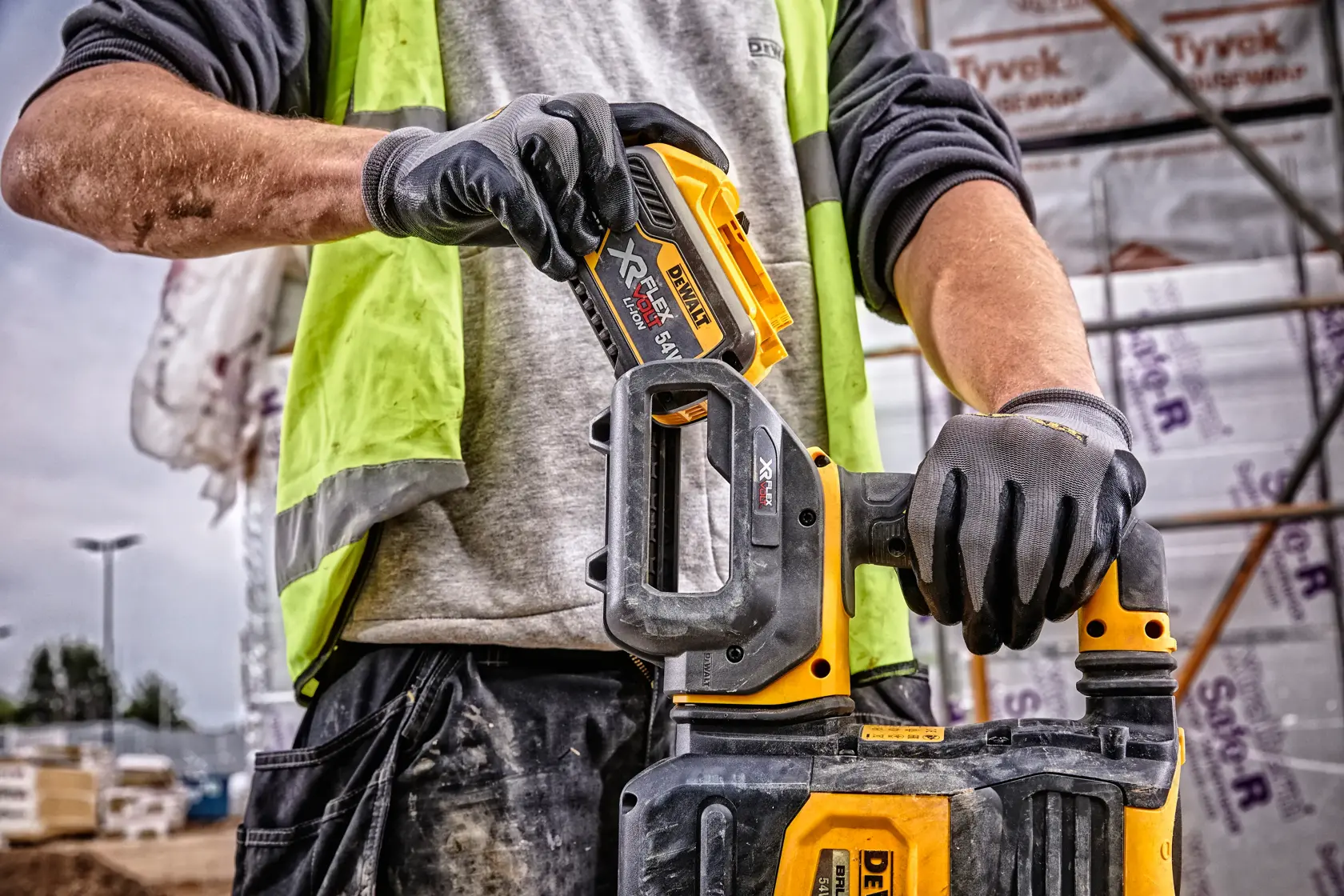 Close-up of a person wearing gloves and a high-visibility vest, inserting a DEWALT DCB548 XR FLEXVOLT battery into a compatible DEWALT power tool at a construction site.