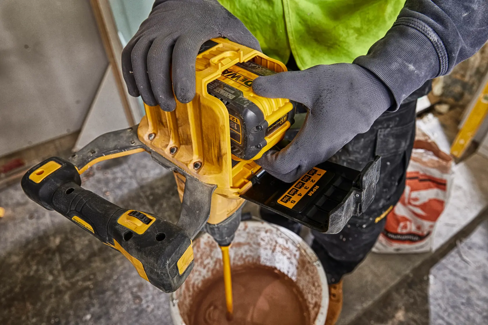 A person wearing gloves uses a yellow DEWALT cordless mixer with a DEWALT DCB548 battery to mix material in a bucket on a construction site.