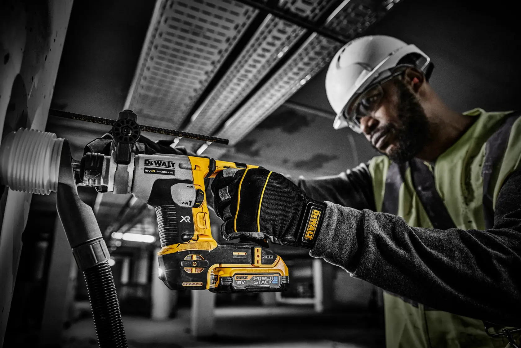 Worker drills into a wall with a DEWALT PowerStack 18V tool, wearing gloves and helmet on a construction site.