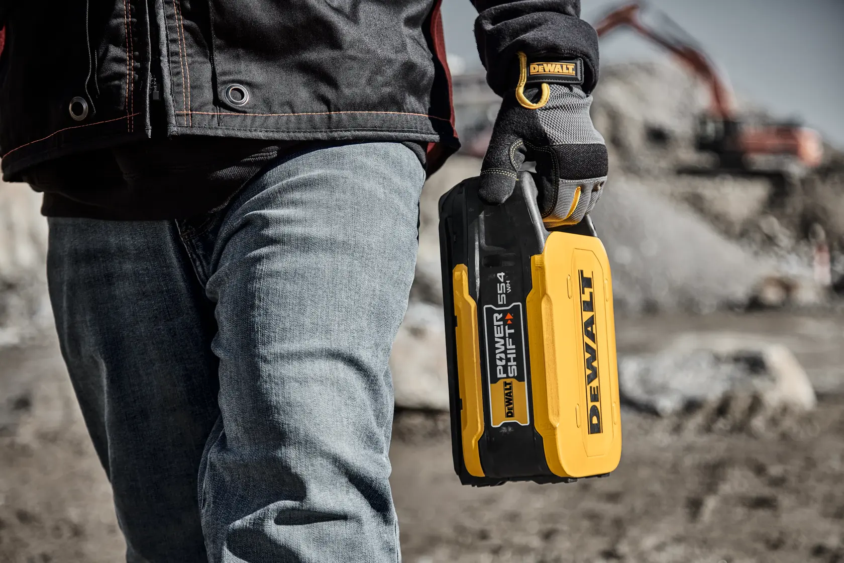 Person holding DEWALT DCBPS0554 battery outdoors at a construction site, wearing gloves and jeans.