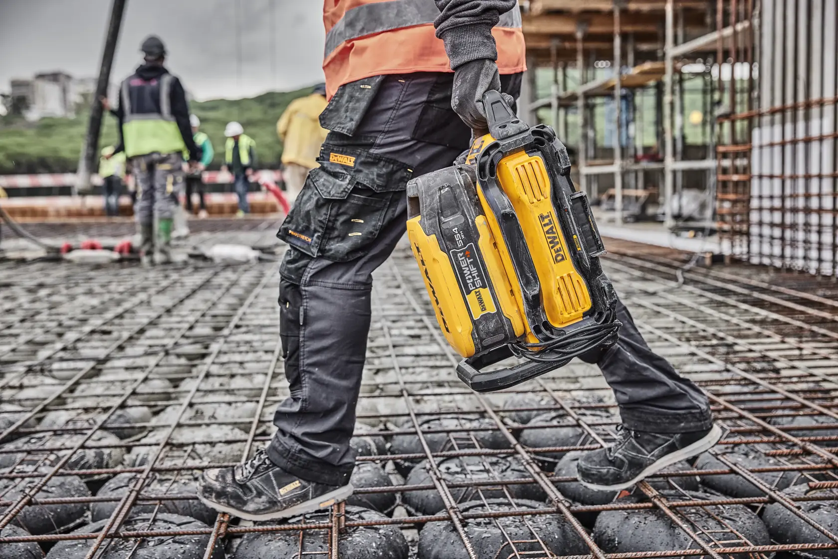Person carrying a DEWALT DCBPS0554 battery on a construction site, wearing safety gear and work boots.