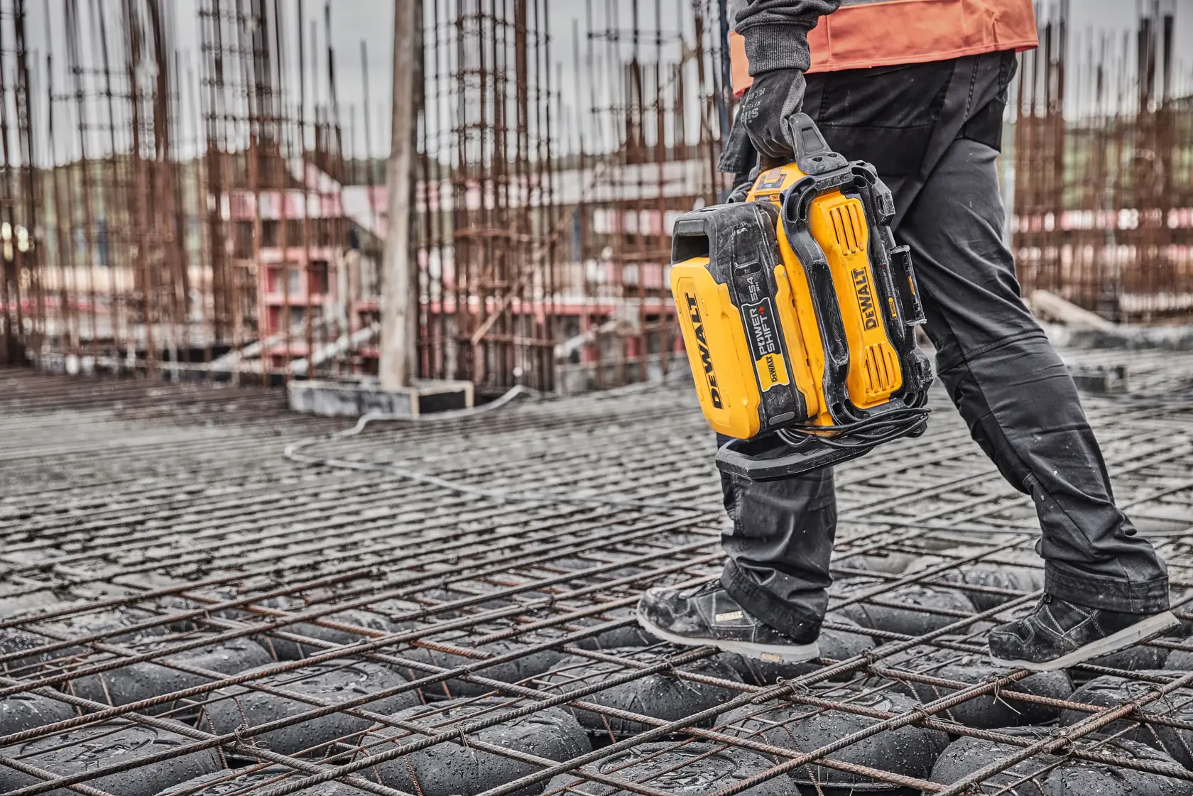 Person carrying a yellow DEWALT battery tool at a construction site with steel reinforcement and unfinished structures.