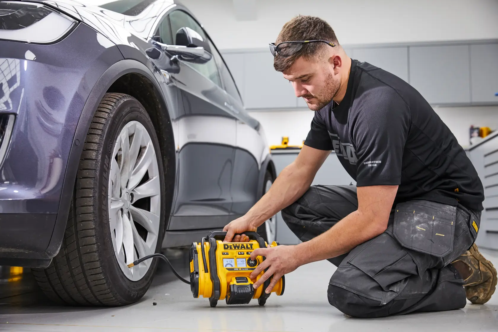 A person kneeling on the floor is using a DEWALT DCC018N portable air compressor to inflate the tire of a parked car inside a garage or workshop. The person's face is blurred for privacy.