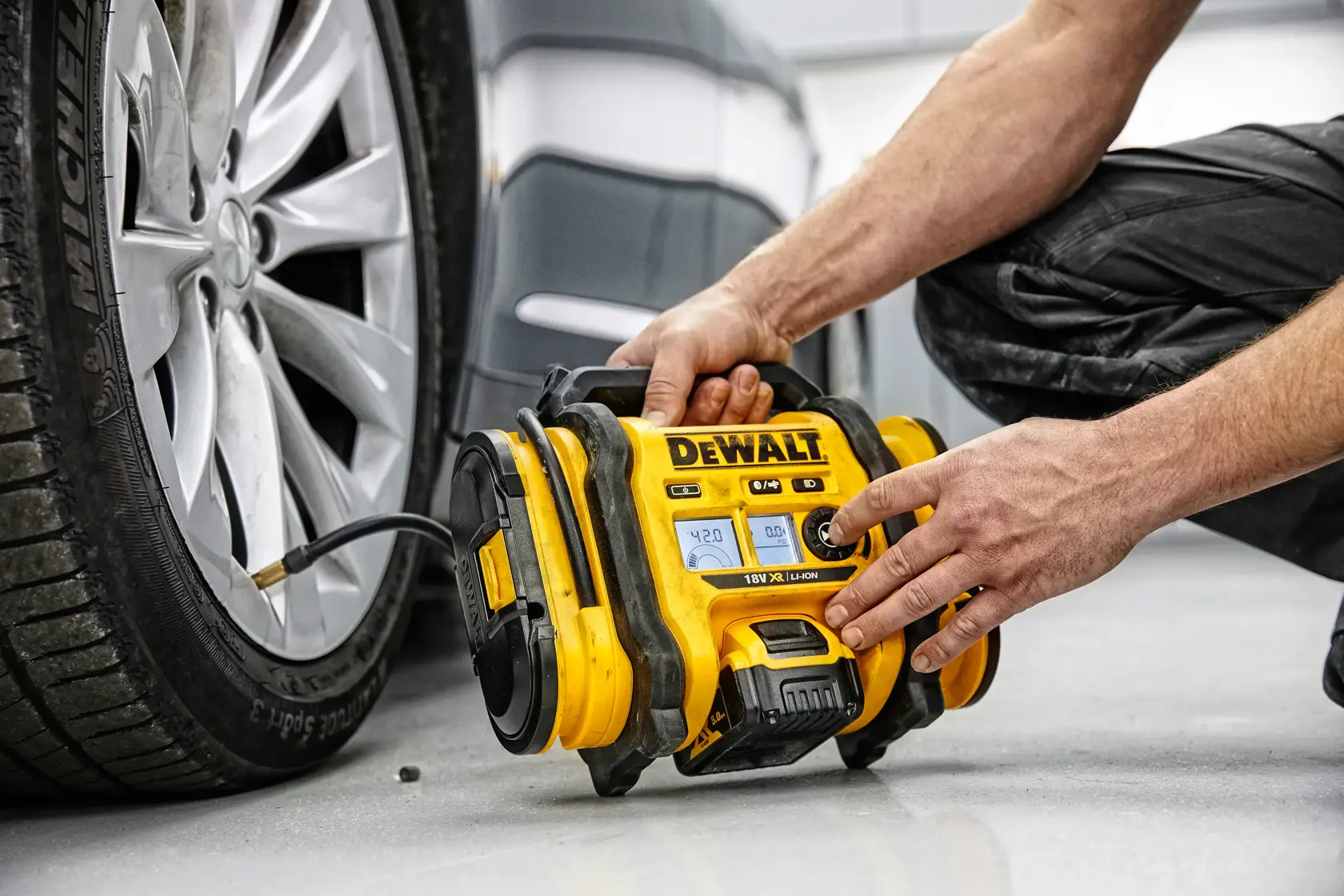 A person using a yellow DEWALT portable air compressor to inflate the tire of a car. The device is connected to the tire valve and placed on the ground next to the vehicle.