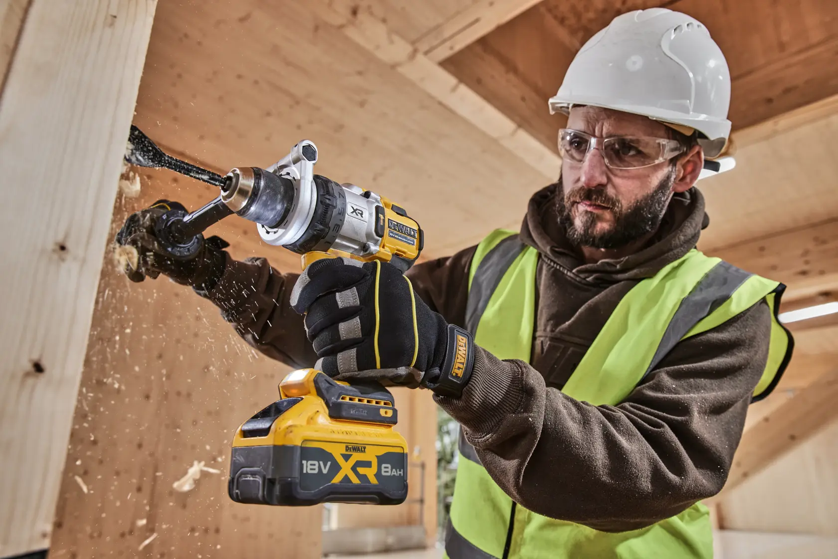 Worker drills wood with DEWALT 18V XR tool, wearing gloves, helmet, and safety vest on construction site.