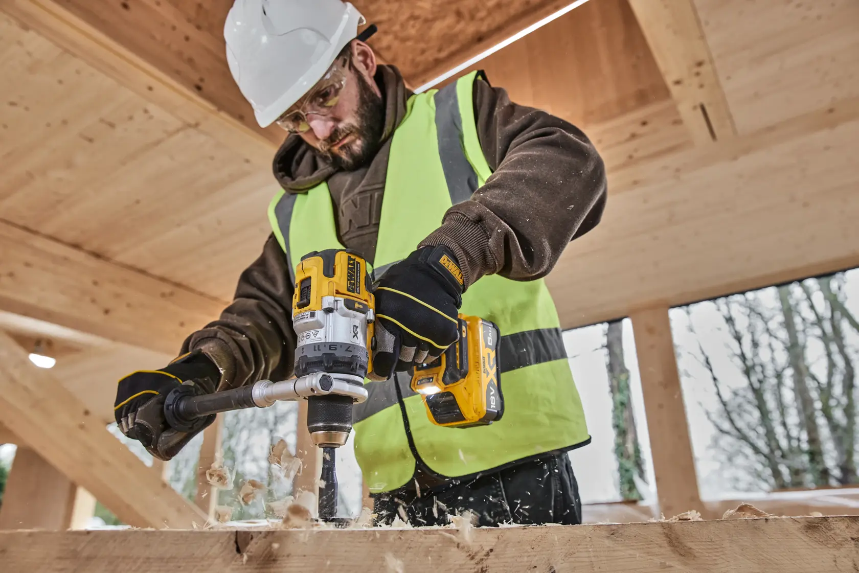 Person uses DEWALT cordless drill to bore wood, wearing safety gear and gloves at a wooden construction site.