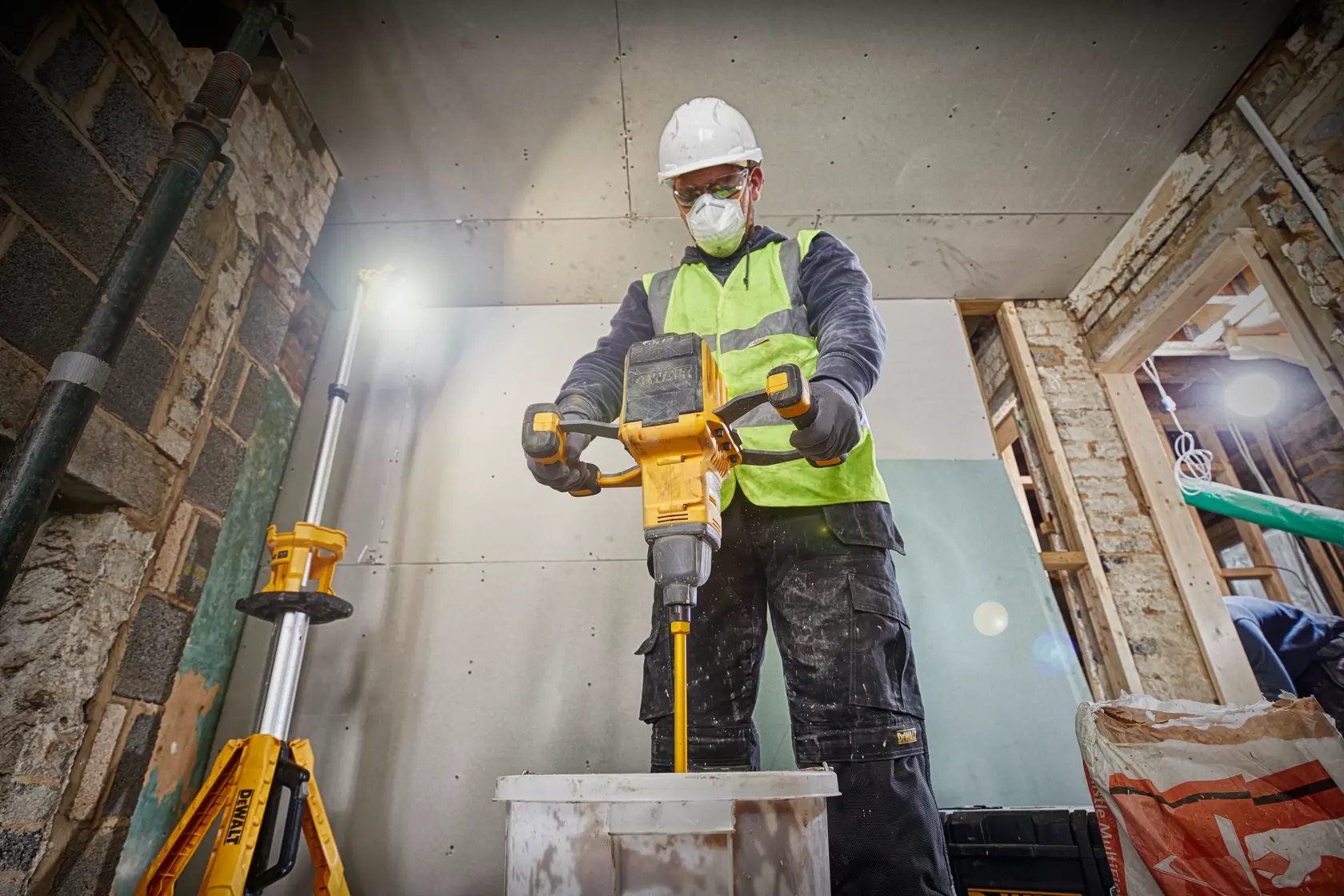 Worker mixes materials with a DEWALT tool indoors, wearing safety gear and a high-visibility vest.