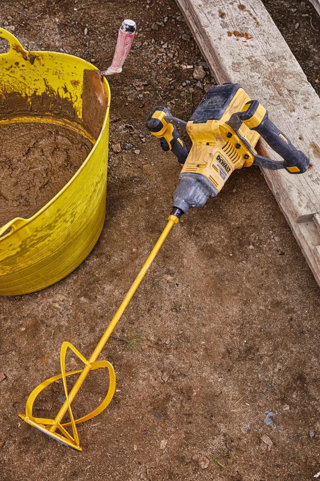 A DEWALT cordless mixer tool with a long mixing paddle attached, placed on a construction site next to a yellow tub containing mixed material.