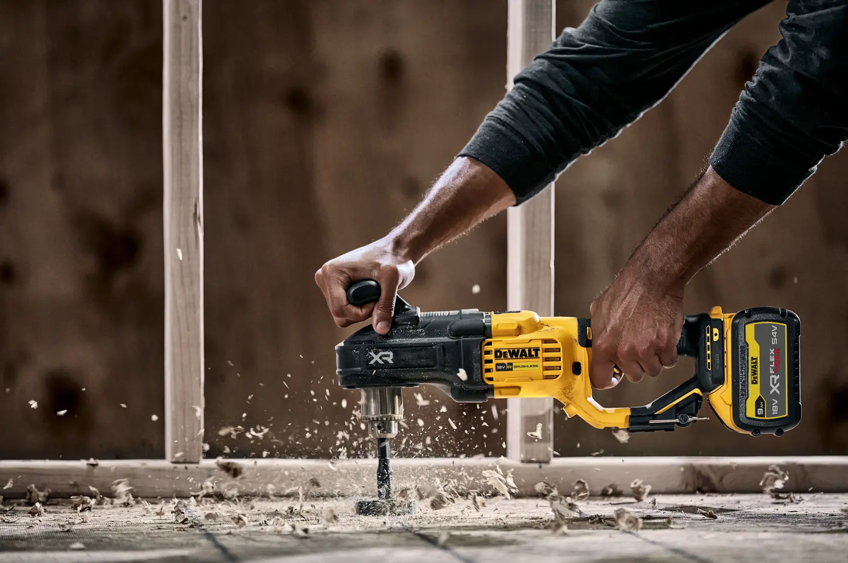 En person bruker en DEWALT DCD444N trådløs drill med sidehåndtak for å bore gjennom tre, noe som skaper trespon og støv. Bygningsramme er synlig i bakgrunnen.