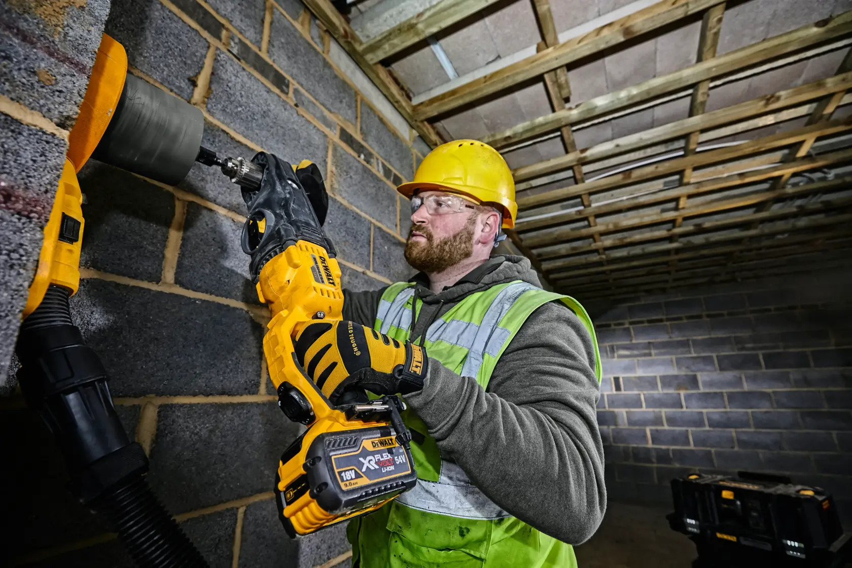 A person wearing a yellow hard hat, safety gloves, and a high-visibility vest is using a DEWALT cordless drill (DCD470N) to bore a hole into a concrete block wall at a construction site. The person's face is blurred.