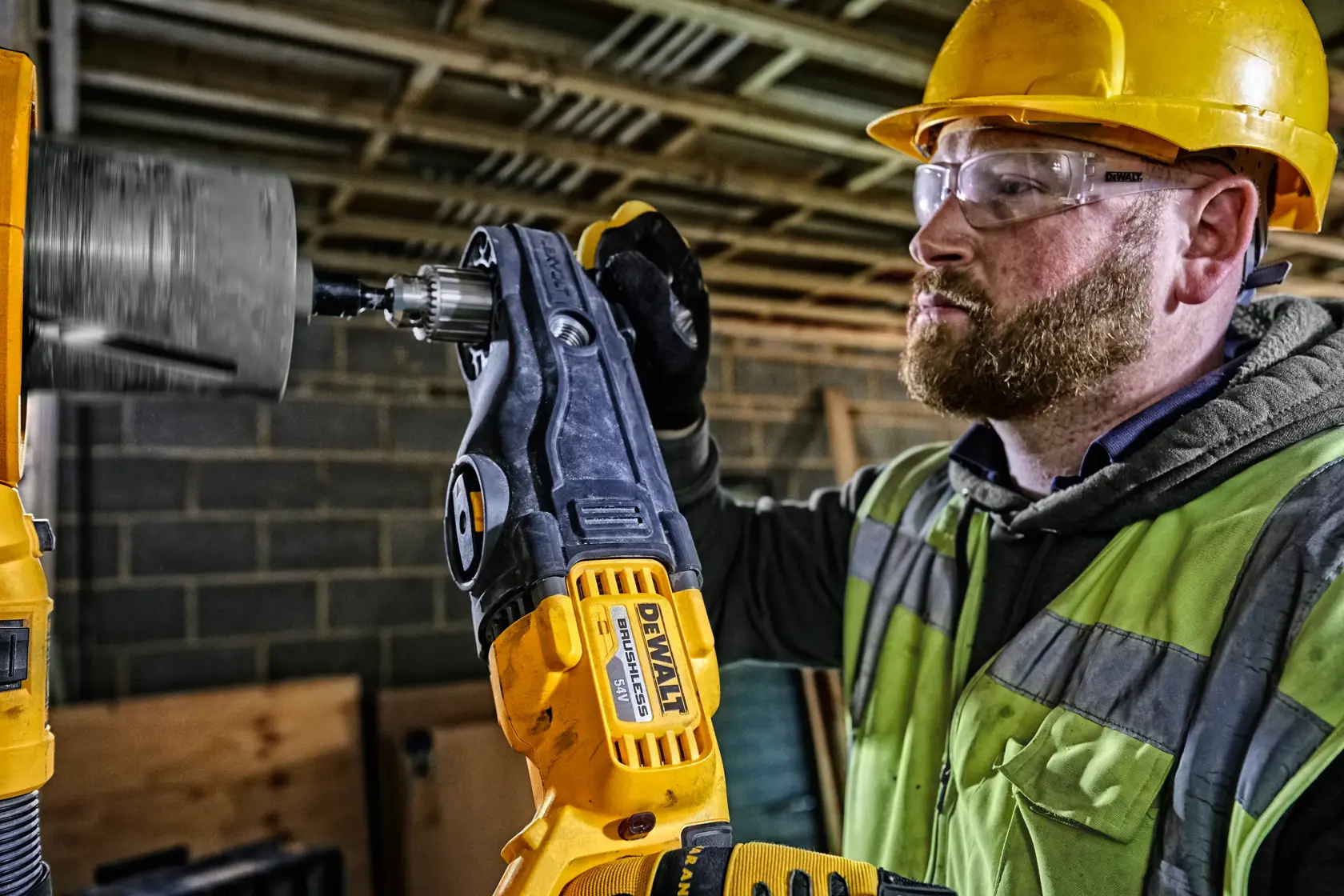 A person wearing a yellow hard hat and safety vest is using a DEWALT DCD470N power drill with an attachment to drill into a surface at a construction site. The person’s face is blurred for privacy.