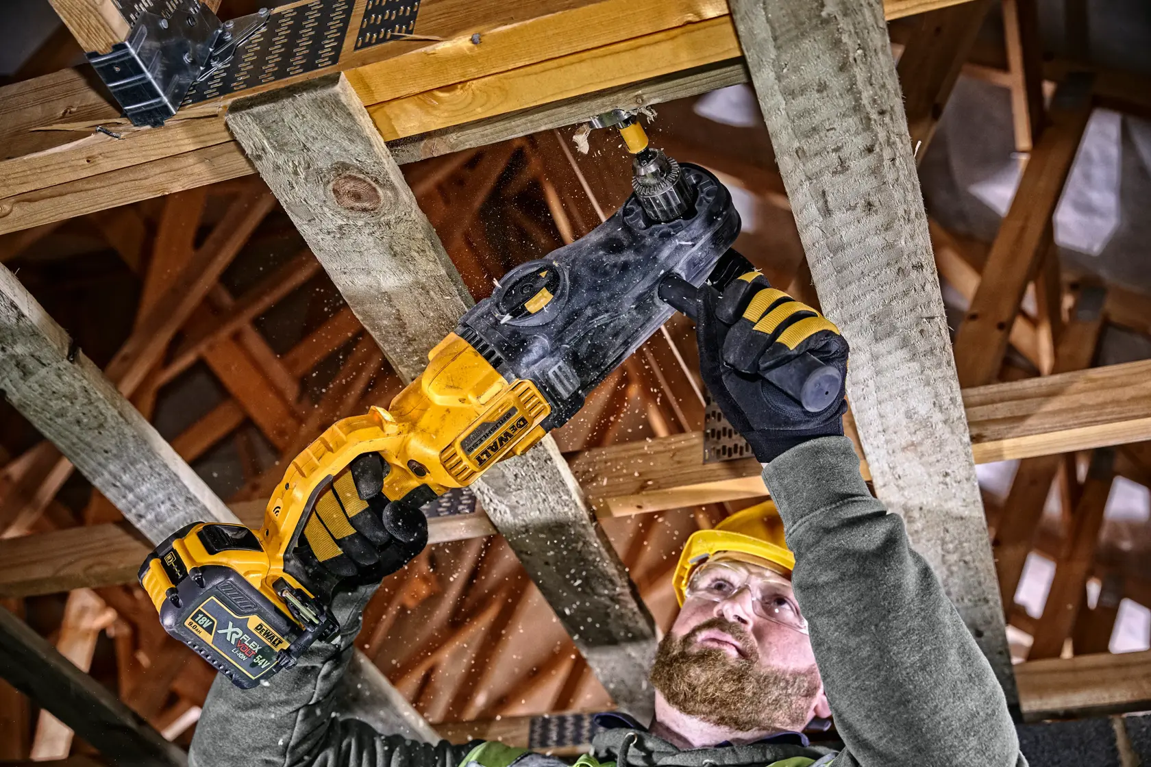 A person using a DEWALT DCD470N cordless drill to drill a hole in a wooden beam as part of construction work. The worker is wearing protective gloves and a safety helmet.