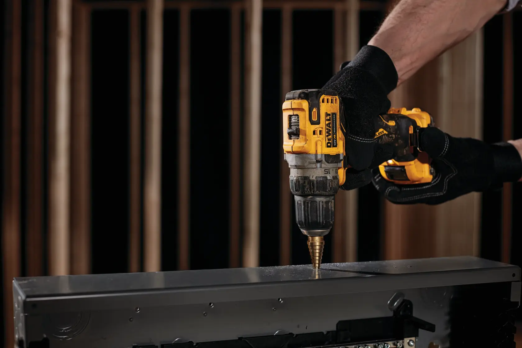 A person wearing black gloves using a yellow DEWALT cordless drill to make a hole in a metal surface.
