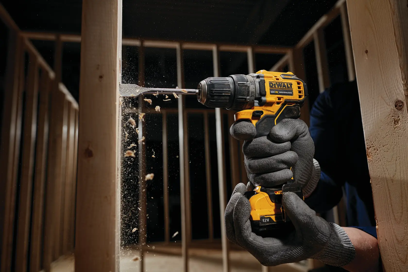A person wearing protective gloves uses a yellow DEWALT cordless drill to drill into a wooden beam inside a framed structure. Wood chips are visible flying from the drilling process.
