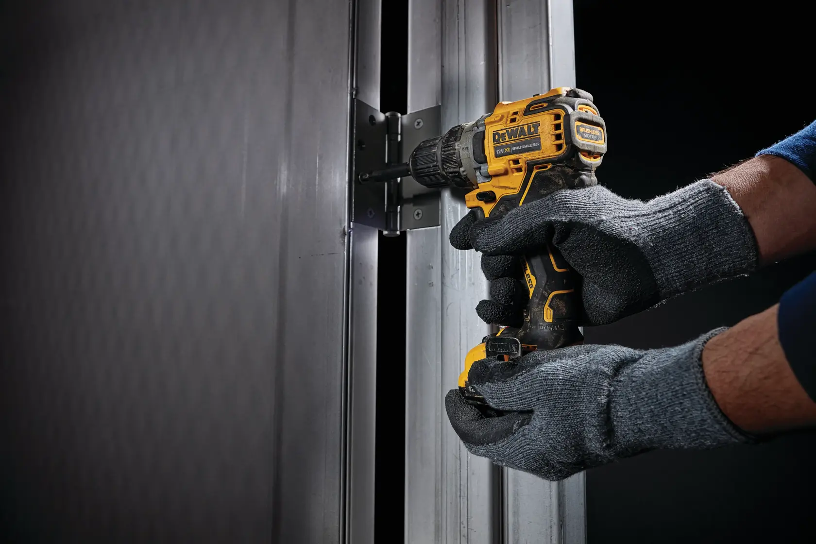 A person wearing gray gloves uses a yellow and black DEWALT cordless drill to secure a hinge on a metal door frame.