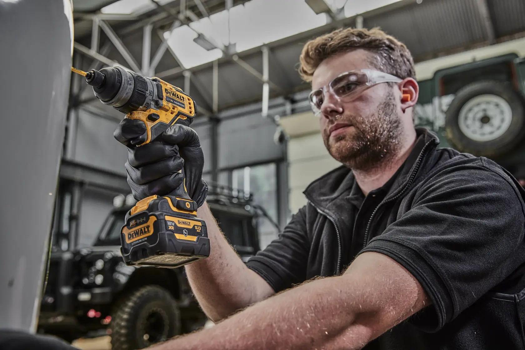 A person with a blurred face is using a DEWALT DCD703NT cordless power drill in a workshop or garage setting. The drill is yellow and black, and the person is wearing black gloves.