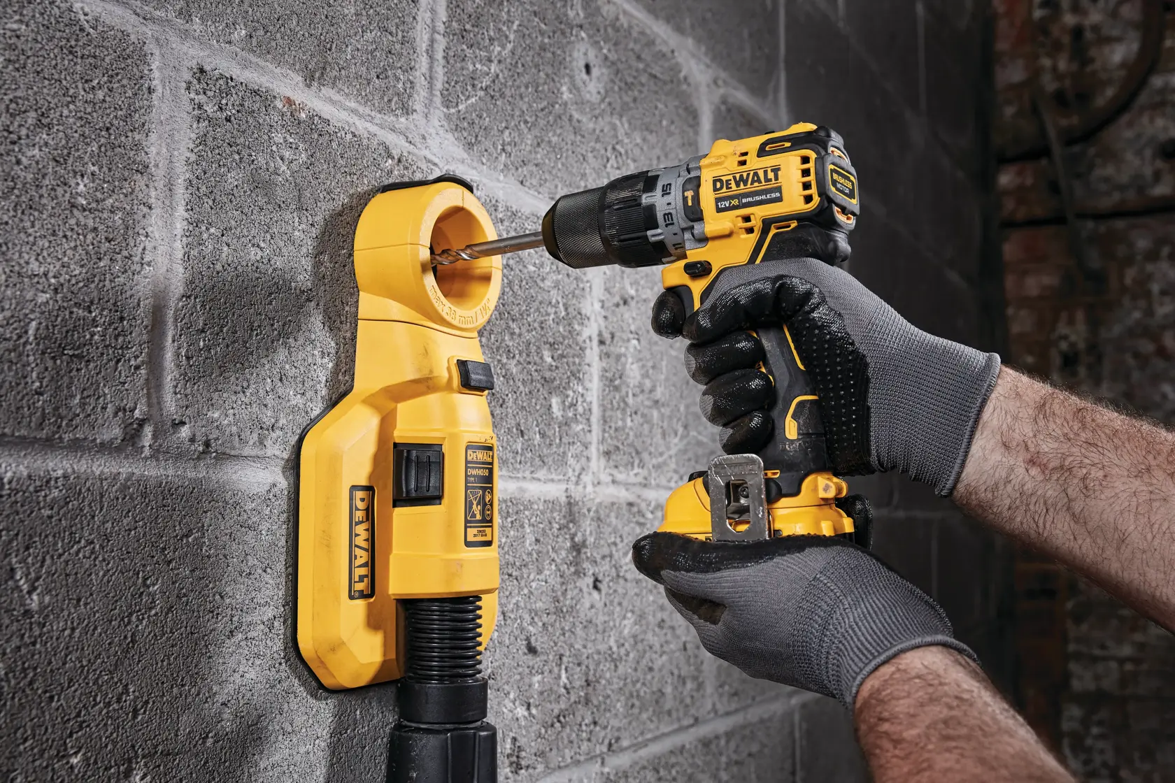 A person wearing gray gloves is using a yellow DEWALT cordless drill to drill into a concrete wall, with a yellow dust extractor attached to collect debris.