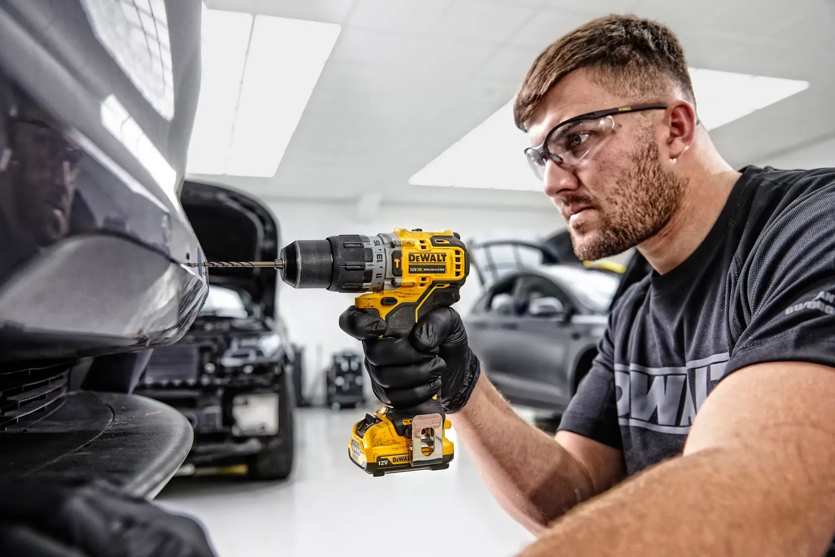 A person wearing black gloves uses a yellow DEWALT DCD706D2 cordless drill to drill into the bumper of a car in an automotive workshop. The person's face is blurred and several cars are visible in the background.