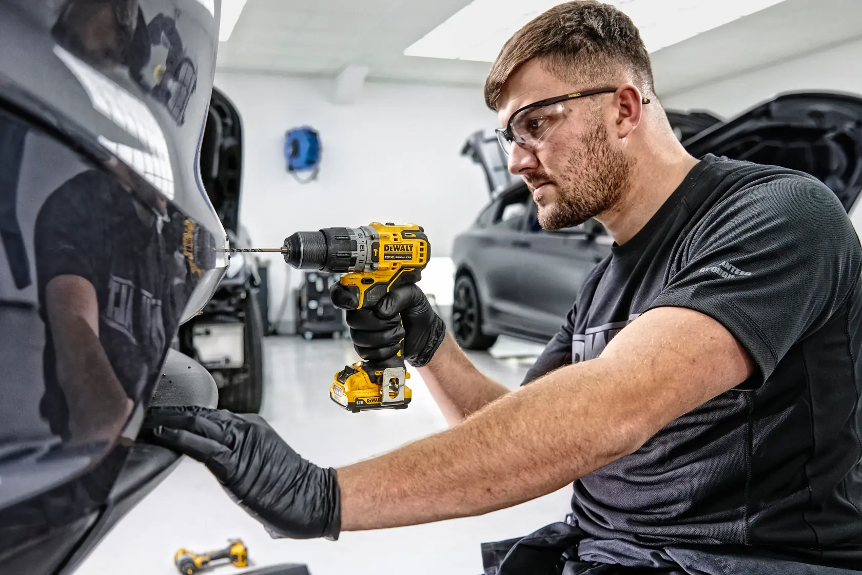 A person wearing black gloves uses a DEWALT cordless drill on the rear bumper of a car in a workshop setting. The person's face is blurred for privacy.
