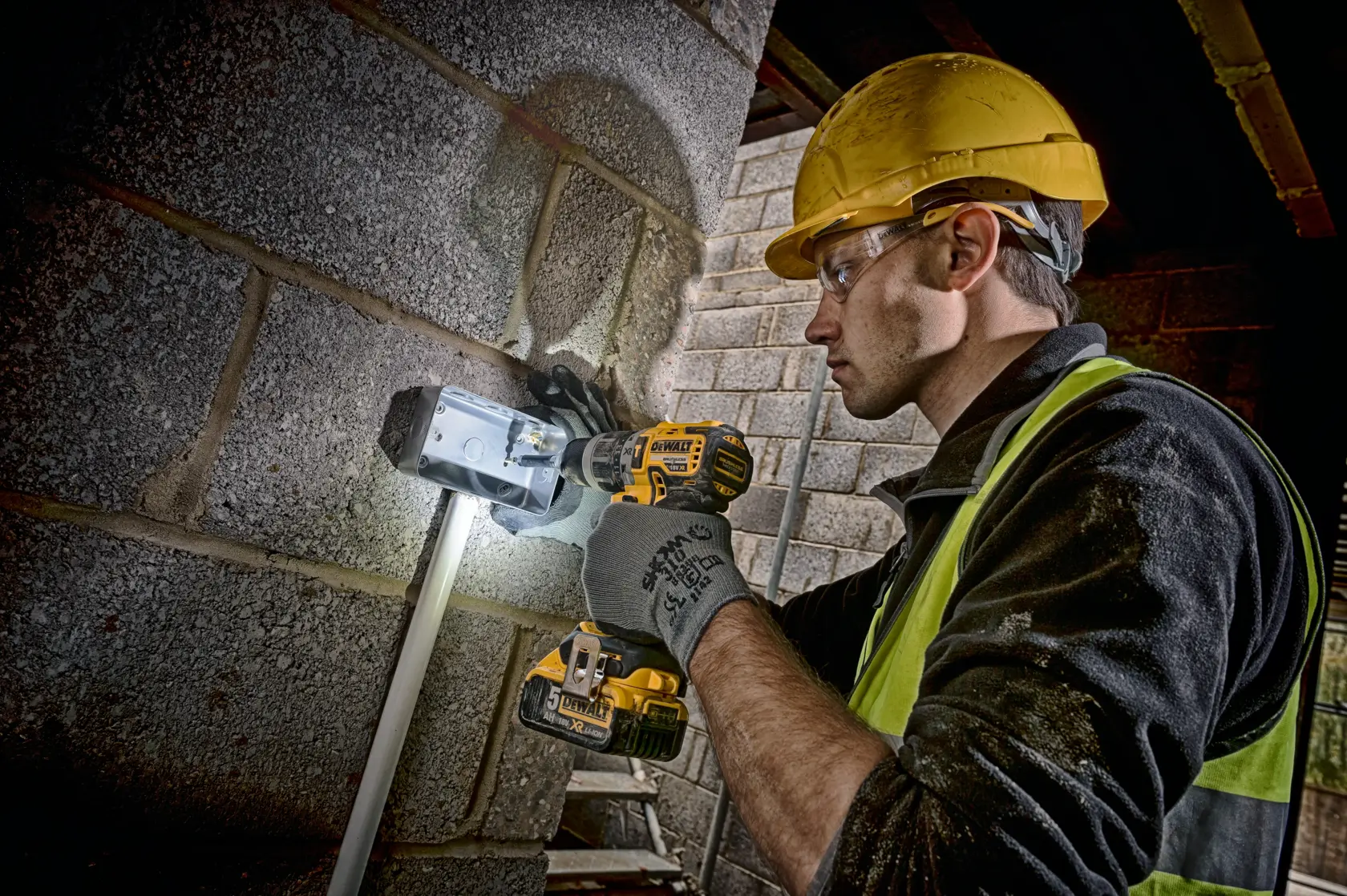 A person wearing a yellow hard hat and safety vest is using a DEWALT DCD796P2 cordless drill to work on a metal electrical box mounted on a brick wall. The person's face is blurred for privacy.