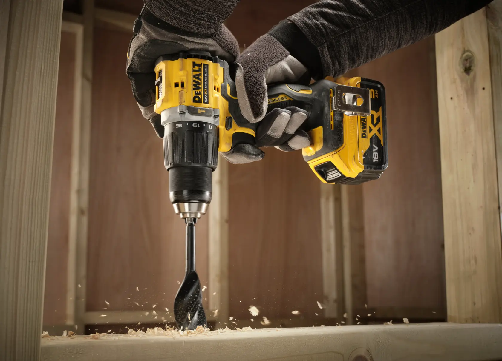 DEWALT cordless drill in use, drilling wood with wood shavings flying, held by gloved hands at a construction site.