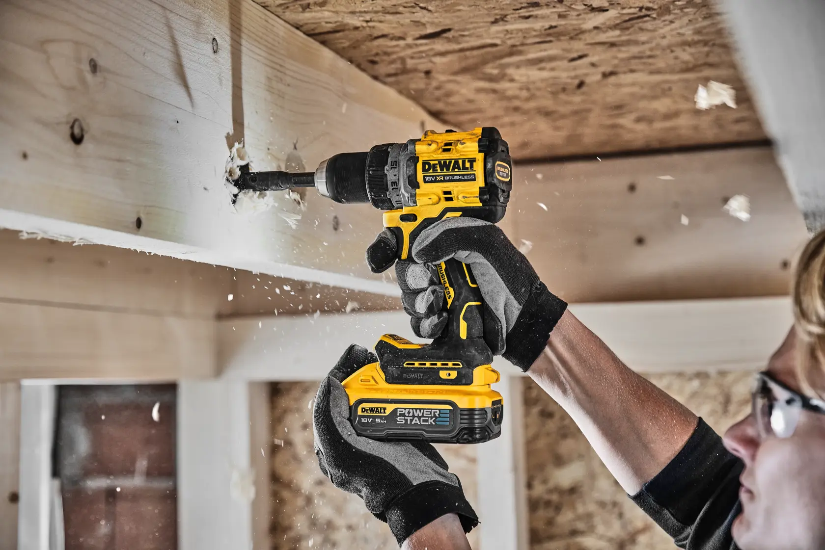 Person drills wood beam with DEWALT cordless drill, wearing gloves and safety glasses in a workshop.