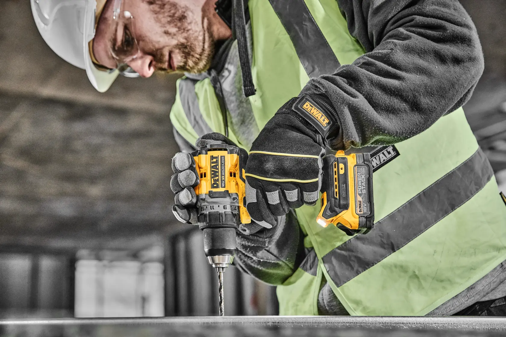 A person wearing safety gear, including a helmet, goggles, gloves, and a high-visibility vest, is using a yellow and black DEWALT cordless power drill at a construction site.