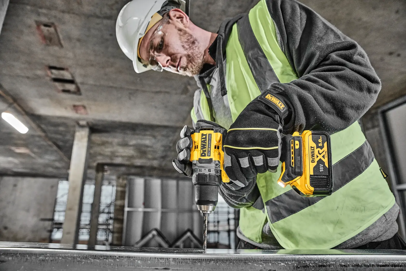 A person wearing safety gear, including gloves, a helmet, and a hi-vis vest, is using a DEWALT DCD800P2T cordless drill to work on a metal surface in an industrial environment.