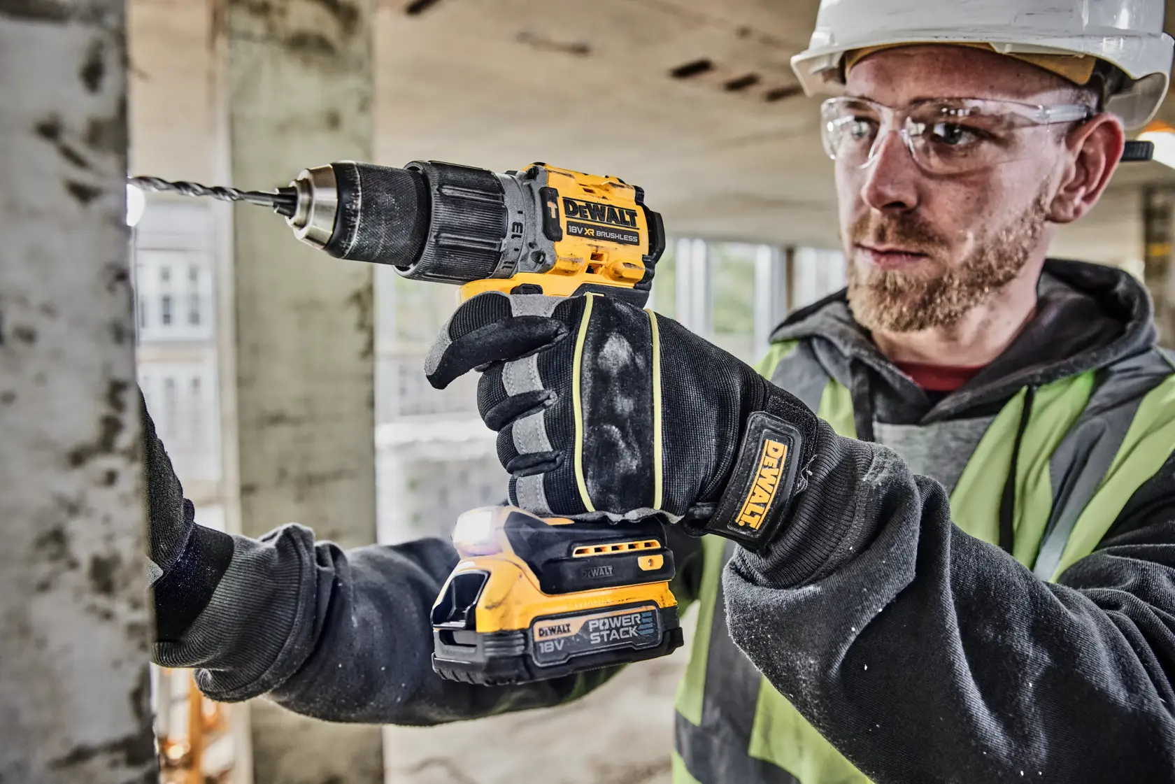 A person wearing safety gloves and a safety vest is using a DEWALT 18V cordless drill to drill into a metal surface at a construction site. The drill is branded DEWALT and fitted with a POWERSTACK battery.