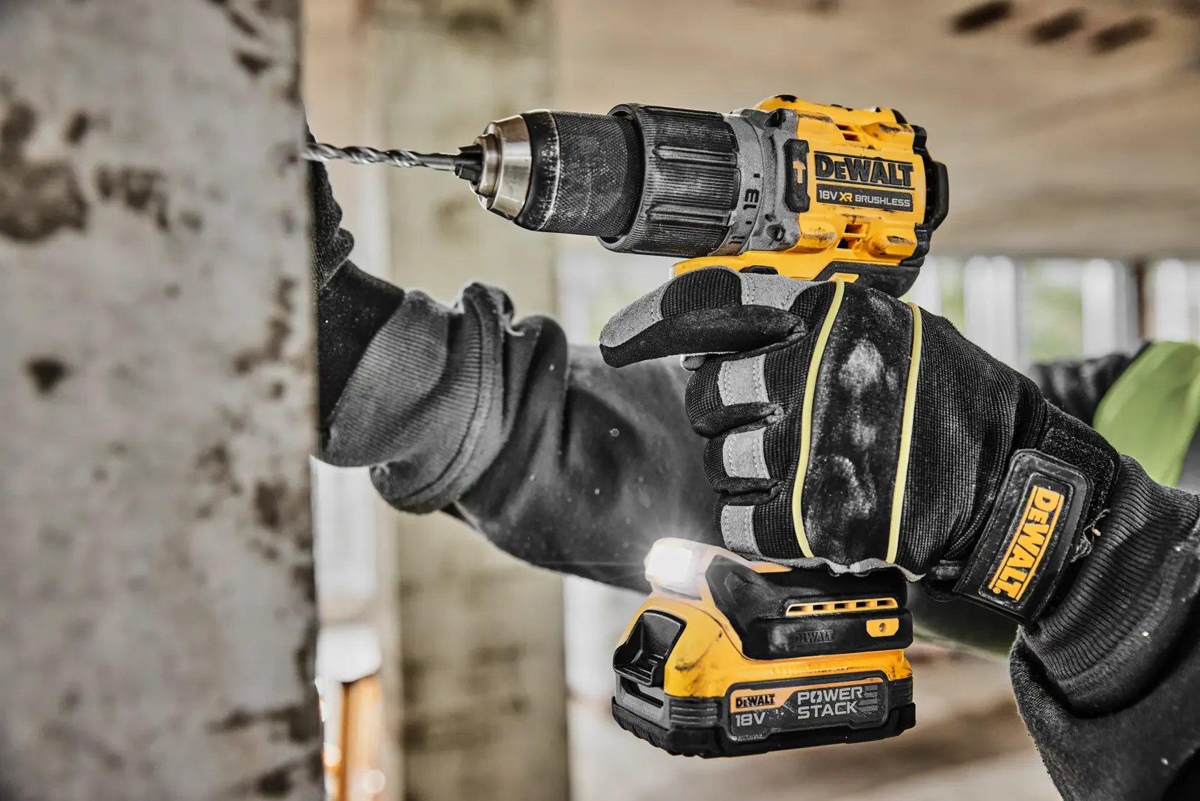 A person wearing black work gloves is using a yellow and black DEWALT cordless drill to drill into a concrete surface indoors.