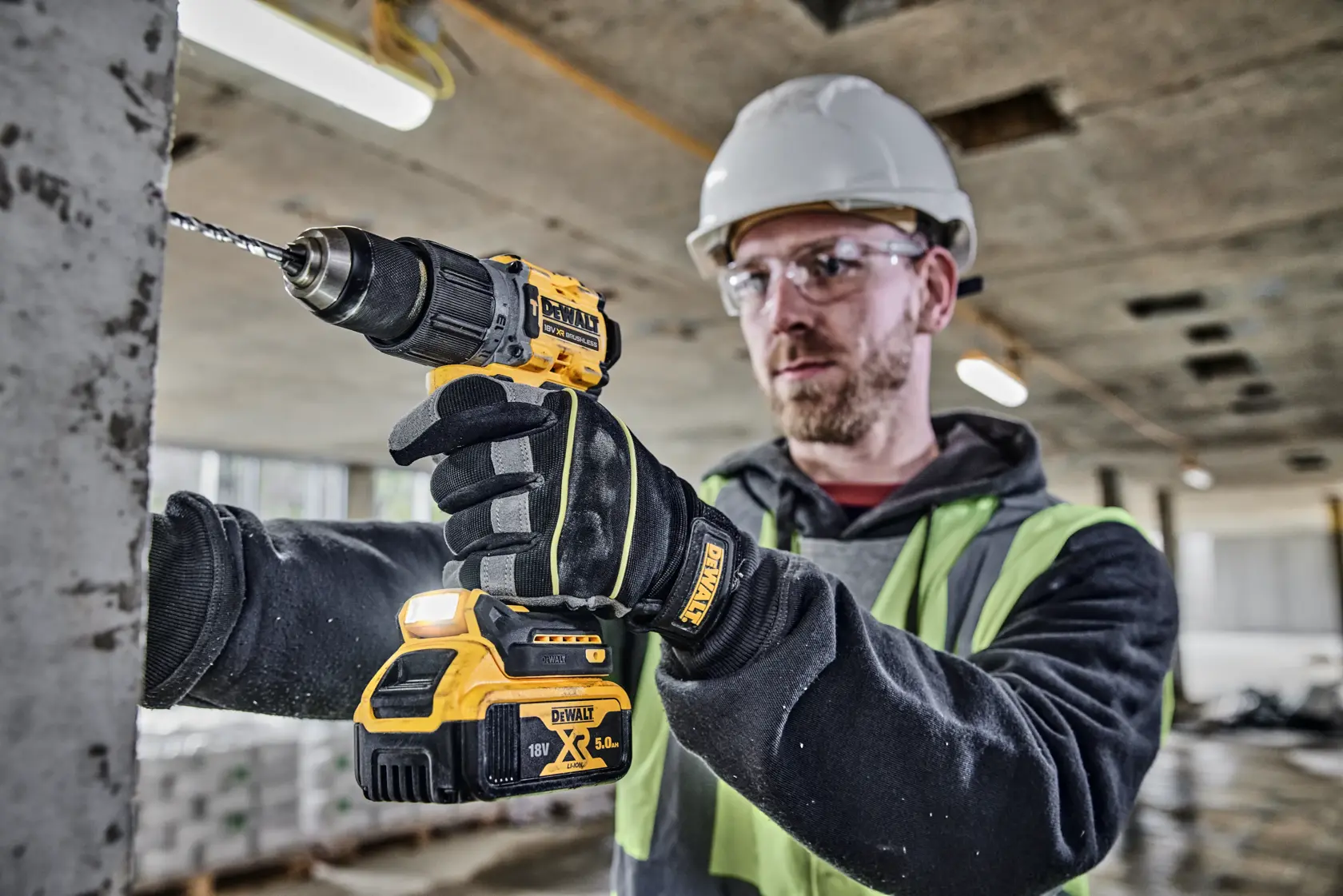 Una persona con guantes, chaleco de seguridad y casco utiliza un taladro inalámbrico DEWALT DCD805P2T para perforar una superficie de hormigón en una obra. El rostro está difuminado por motivos de privacidad.