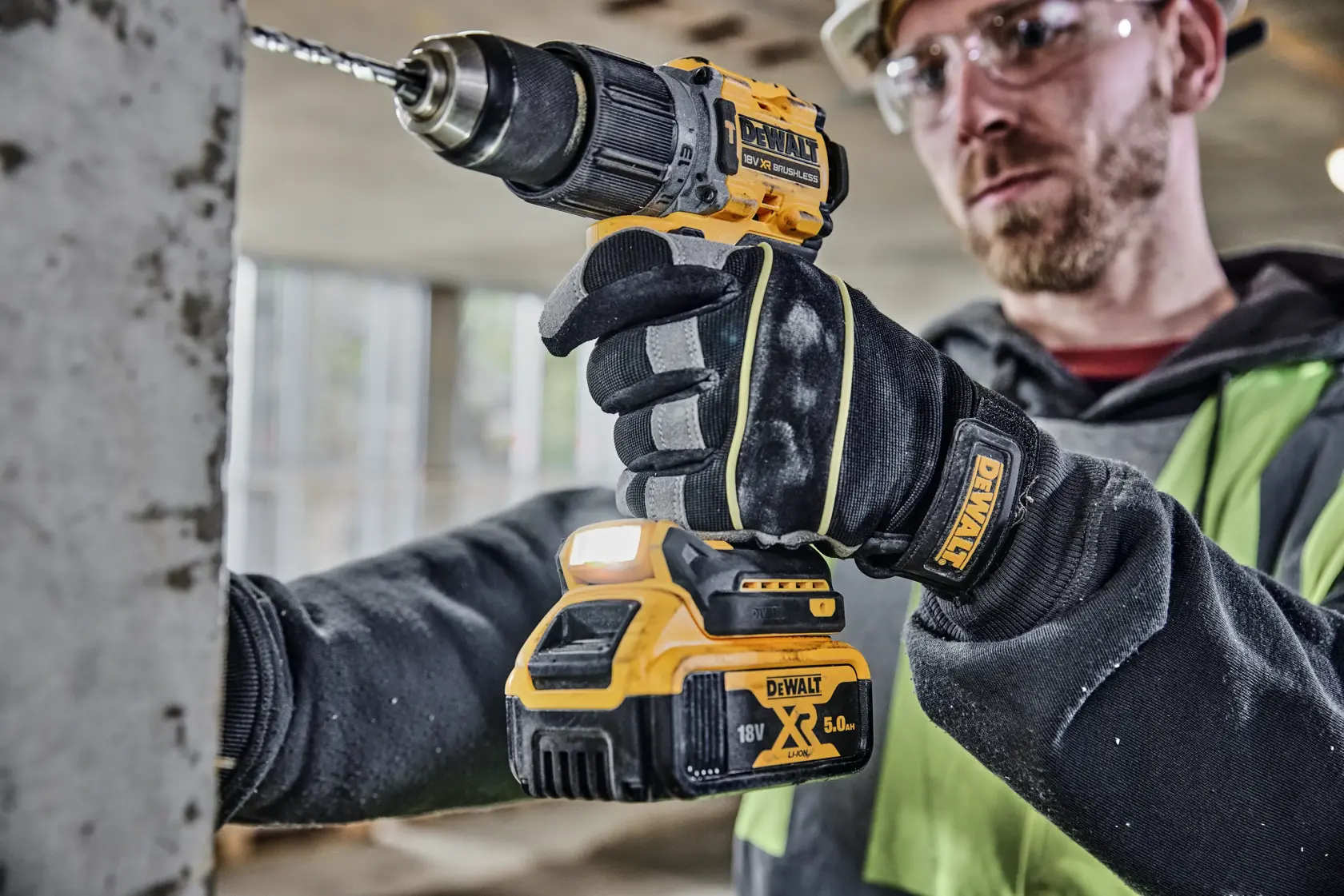 Una persona con guantes de la marca DEWALT está usando un taladro atornillador inalámbrico DEWALT 18V XR (modelo DCD805P2T) para perforar una superficie de concreto en un entorno de construcción interior. El rostro de la persona está desenfocado.