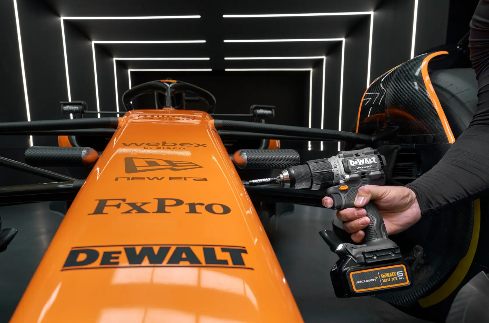 A DEWALT cordless drill is used on an orange McLaren race car, both branded with sponsor logos in a modern garage.
