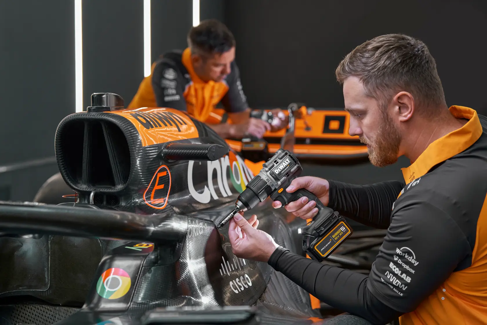 Person uses a DEWALT power drill on an orange race car. Faces are blurred and team shirts are visible.